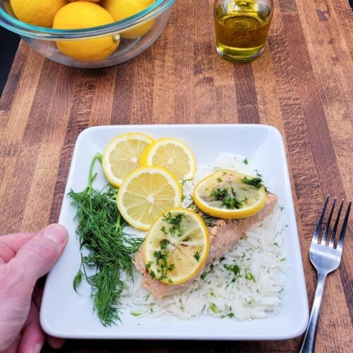 A hand holding a plate of oven baked salmon with lemon and dill.
