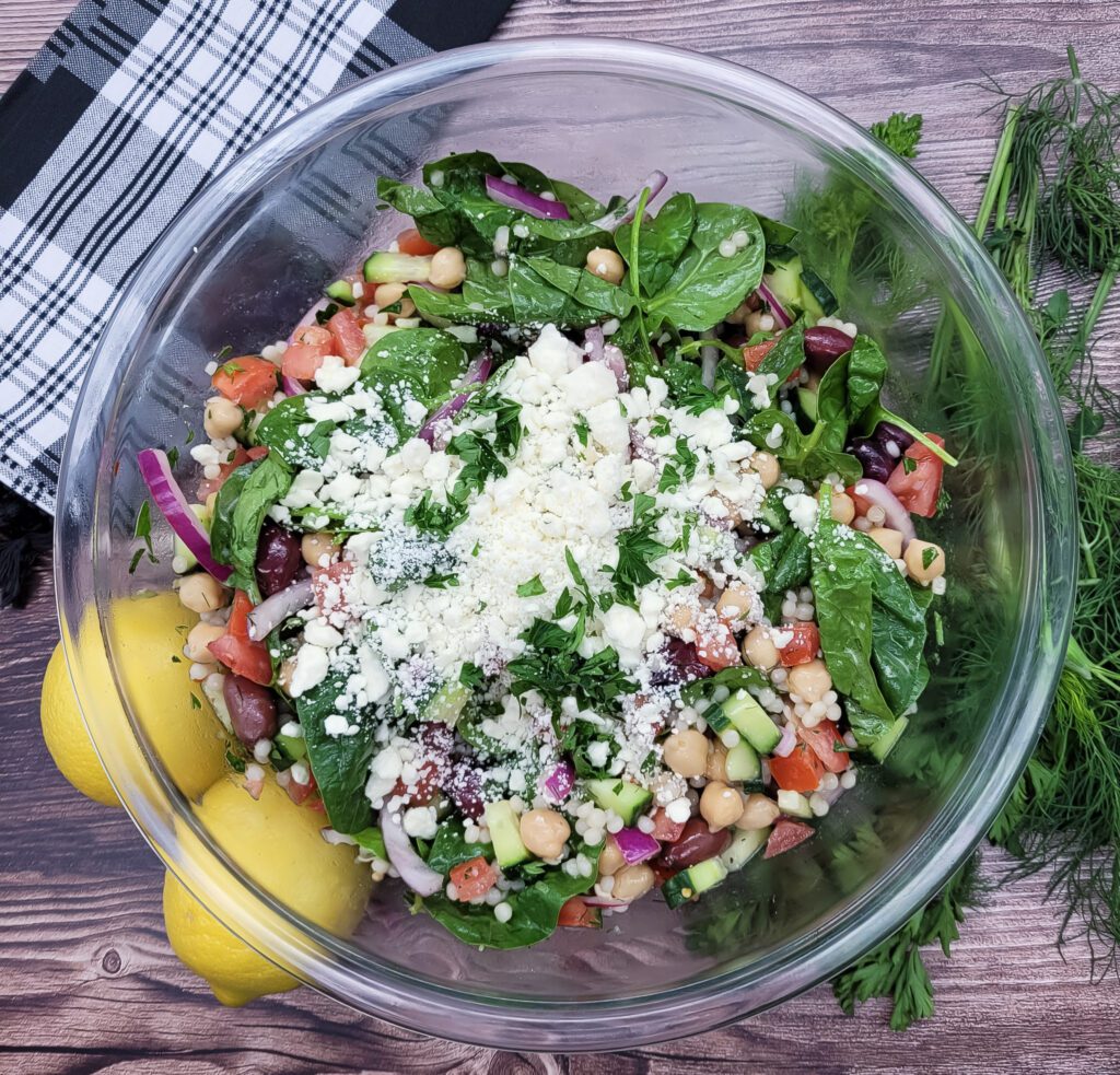 An overhead photo of Mediterranean Tomato Cucumber Salad in a clear bowl, accessorized with a black and white napkin, two lemons, and dill and parsley sprigs