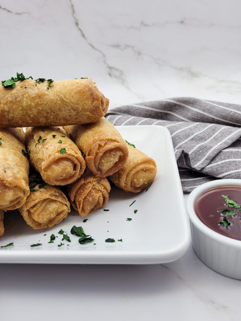 A stack of frozen air fryer egg rolls on a square white plate with a cup of dipping sauce and a gray striped napkin
