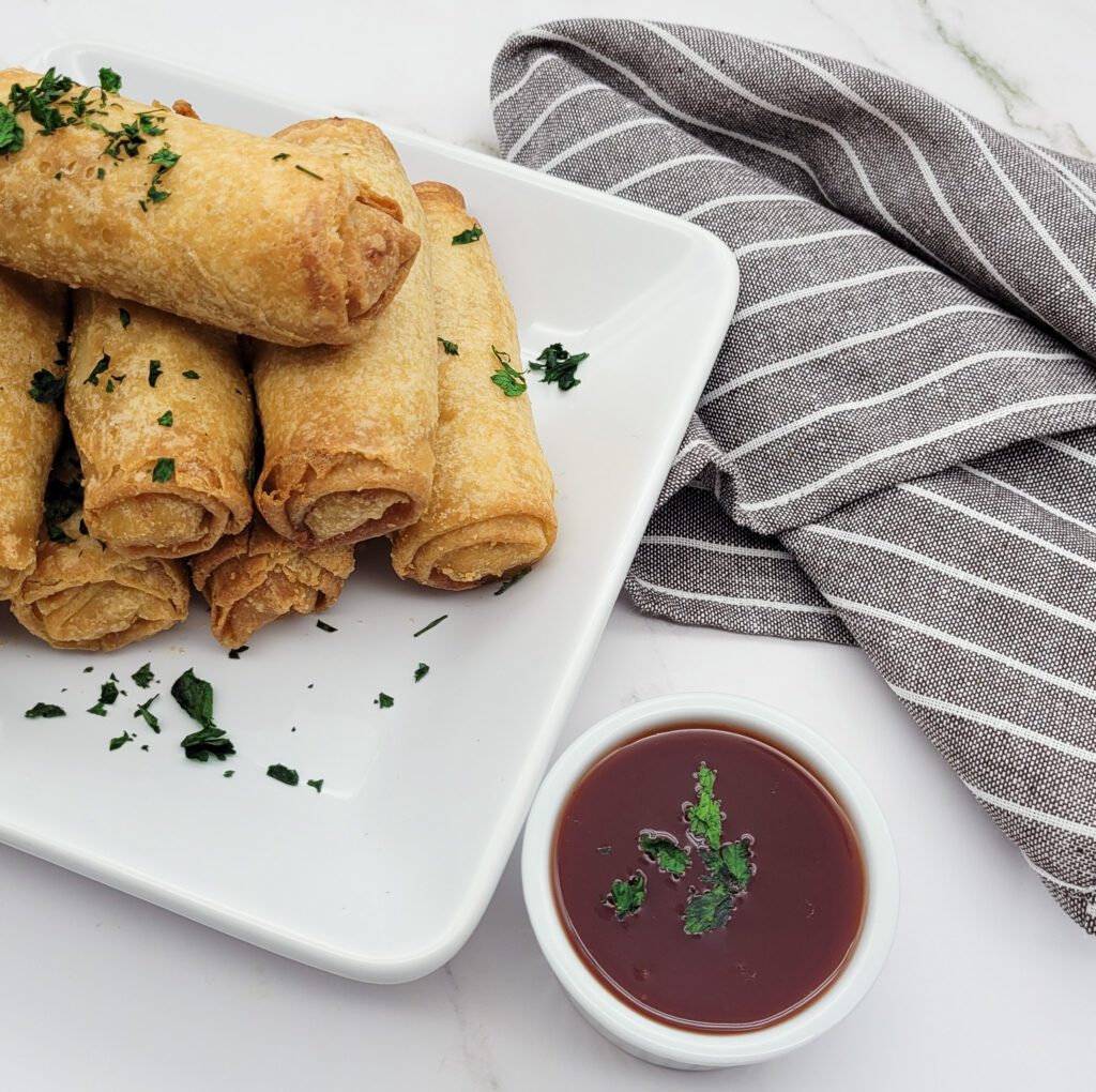 A square plate of air fried frozen egg rolls with a cup of dipping sauce and a gray striped napkin
